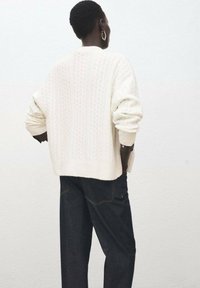 Suéter crema de punto con trenzas, de corte holgado y hombros caídos, combinado con pantalones de mezclilla oscuros con costuras amarillas.