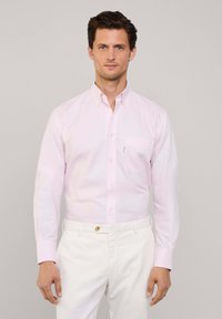 Camicia a maniche lunghe color rosa chiaro realizzata in cotone, con tasca sul petto sinistro, indossata con pantaloni bianchi. Texture liscia, design classico.