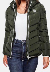 Olivgrön pufferjacka med huva, vit dragkedja och logotypdetalj på bröstet. Den har en quiltad textur och bärs över en vit topp och jeans.