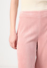 Roze kordura hlače s vertikalnim rebrastim uzorkom, imaju široki pojas i bočne džepove. Mekana tekstura i opušteni kroj.