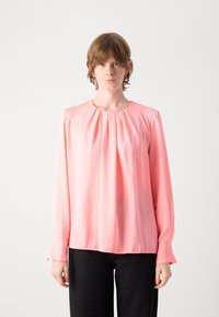 Langärmlige Bluse aus zartrosafarbenem Stoff, mit rundem Ausschnitt, plissierter Front und geknöpften Manschetten, kombiniert mit dunklen Jeans.