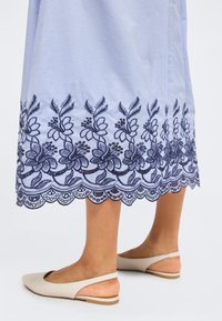 Μπλε φούστα με σκούρα floral κέντημα κατά μήκος της κυματιστής ραφής, φορεμένη με μπεζ slingback μπαλαρίνες σε απλό φόντο.