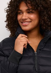 Eine lächelnde Frau mit lockigem Haar zieht eine schwarze Steppjacke mit hervorstehenden Bündchen und Reißverschlussdetails an.
