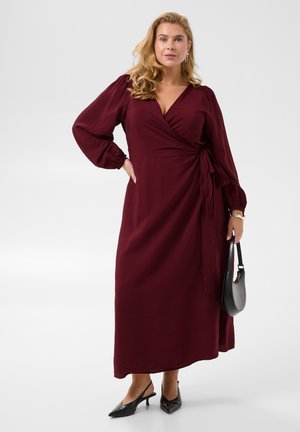Femme aux cheveux blonds porte une longue robe portefeuille bordeaux à manches bouffantes, des escarpins slingback noirs, et un sac à main noir.