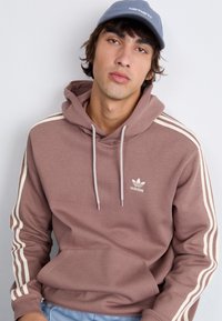 Rudos spalvos džemperis su baltais dryžiais ant rankovių ir išsiuvinėtu Adidas logotipu ant krūtinės. Minkšta medžiaga ir kišenė kengūros stiliumi.