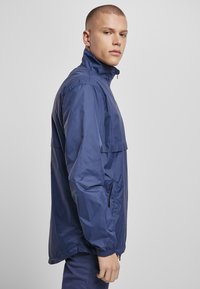Marineblå vindjakke med høj krave, elastiske manchetter og sidelommer. Har en let, glat tekstur og en afslappet pasform.
