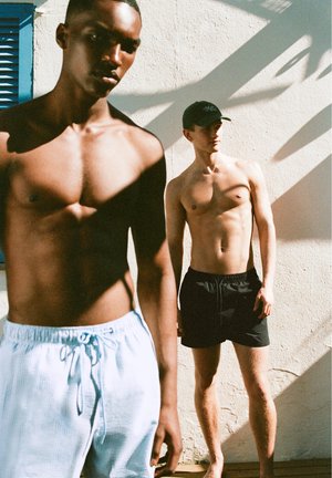 To skjorteløse mænd står op ad en solbeskinnet væg; den ene har lyseblå shorts i forgrunden, den anden har sorte shorts og en kasket i baggrunden.