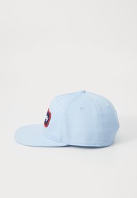 Casquette de baseball bleu clair avec une visière plate, comportant un patch logo brodé rouge et marine à l'avant et des trous de ventilation sur la calotte.