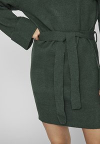 Robe tricotée vert foncé à manches longues avec une ceinture assortie nouée à la taille, montrant une partie de la main et des jambes d'une personne.
