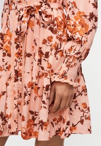 Détail des manches et de la taille d’une robe rose avec un motif floral orange, boutons recouverts de tissu, poignets froncés et ceinture nouée à la taille.