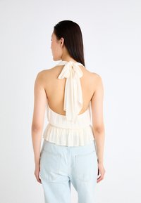 Kremfarget halterneck-top med åpen rygg, knyttet sløyfe i nakken og lagdelt volangkant; brukt med lyseblå høy midje jeans.
