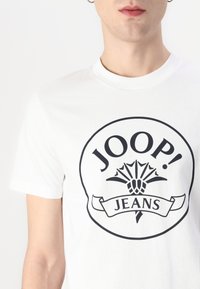 Bílý bavlněný tričko s černým kruhovým logem s texty "JOOP!" a "JEANS", zdobené dekoračním prvkem a detailem stužky.
