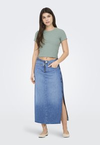 Crop top rayé vert avec un ourlet festonné, associé à une longue jupe en denim bleu avec une fente sur le côté et un ourlet effiloché. Des chaussures plates neutres sont portées.