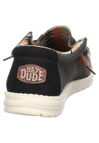 Scarpa slip-on nera in tela con superiore grigio testurizzato, suola bianca e foderatura colorata. Presenta un logo "Hey Dude" sul retro.