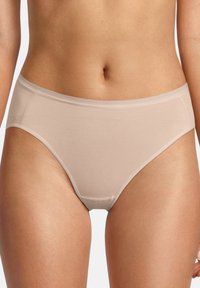 Donna che indossa semplici mutande di cotone beige a vita alta, mostrata dalla vita alle cosce superiori su uno sfondo chiaro e uniforme.