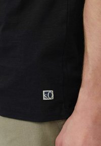 Black short-sleeve shirt featuring a small white and blue branded tag near the hem. Fabric appears soft with a slight texture.