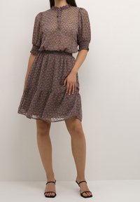 Žena v šaty do kolen s vzorem a nafouknutými rukávy, s elastickým pasem, doplněné černými sandály na podpatku s řemínky.