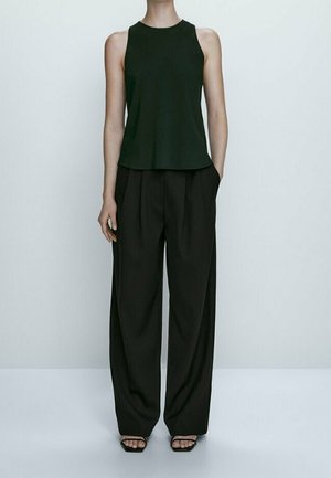 Frau steht und trägt ein ärmelloses schwarzes Oberteil, schwarze Hosen mit weitem Bein und hat die Hände in den Taschen, dazu schwarze offene Sandalen, vor einem einfarbigen Hintergrund.