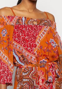 Kvinna i en axelbandslös klänning med orange, röd och blå blommiga och paisley-mönster, knuten med ett matchande tygbälte.