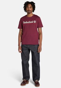 Mörkröd bomullströja med Timberland-logga, ihopparad med mörkblå jeans och bruna snörskor med vita sulor och trädetaljer.