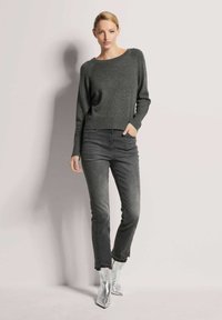 Grauer Strickpullover mit Rundhalsausschnitt, kombiniert mit ausgebleichten schwarzen Skinny-Jeans. Das Model trägt glänzende silberne Stiefeletten. Schlicher Hintergrund.