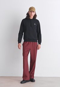 Sweatshirt à capuche noir avec une poche kangourou à l'avant et le logo Lacoste, associé à un pantalon plissé bordeaux et des baskets noires.