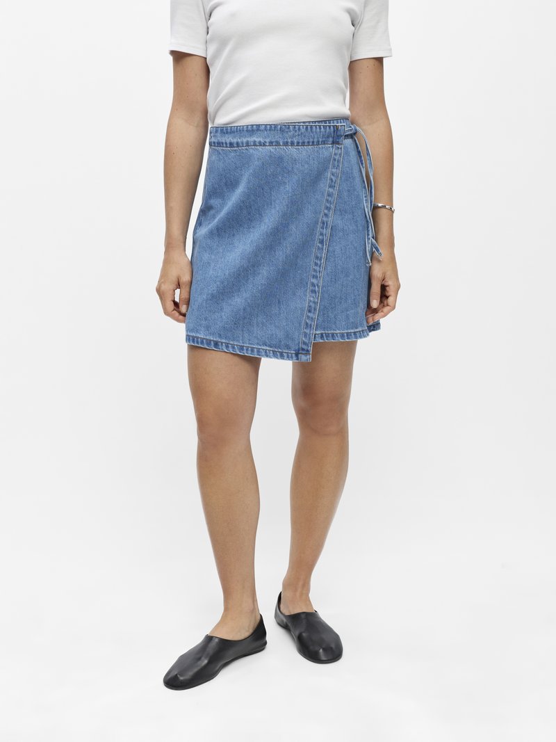 Femme portant un haut blanc à manches courtes, une jupe portefeuille en jean bleu avec des liens sur le côté, des ballerines noires et un bracelet argenté au poignet droit.