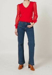 Pull rouge à col en V avec manches bouffantes transparentes à motifs, associé à un jean évasé bleu foncé et des bottines à talons marron.