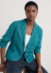 Teal linen blazer with two front pockets, notched lapels, and rolled-up sleeves, featuring a single-button closure. Paired with a white top.