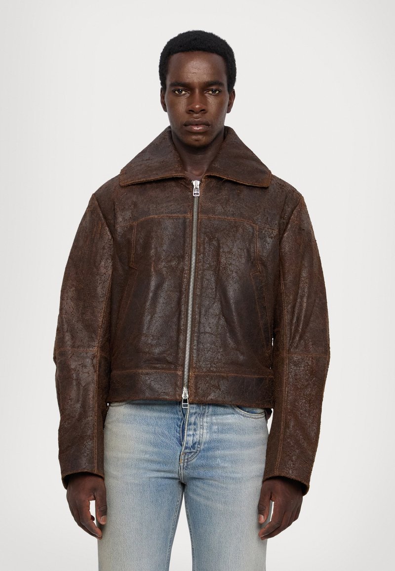 Brown leather jacket with a wide collar, zip closure, and prominent seams. Features front pockets and a cropped silhouette. Worn over light blue jeans.