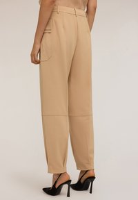 Pantalon taille haute beige avec une coupe ajustée, dotée de poches latérales et d'une texture lisse, associé à des sandales à talons hauts noires.