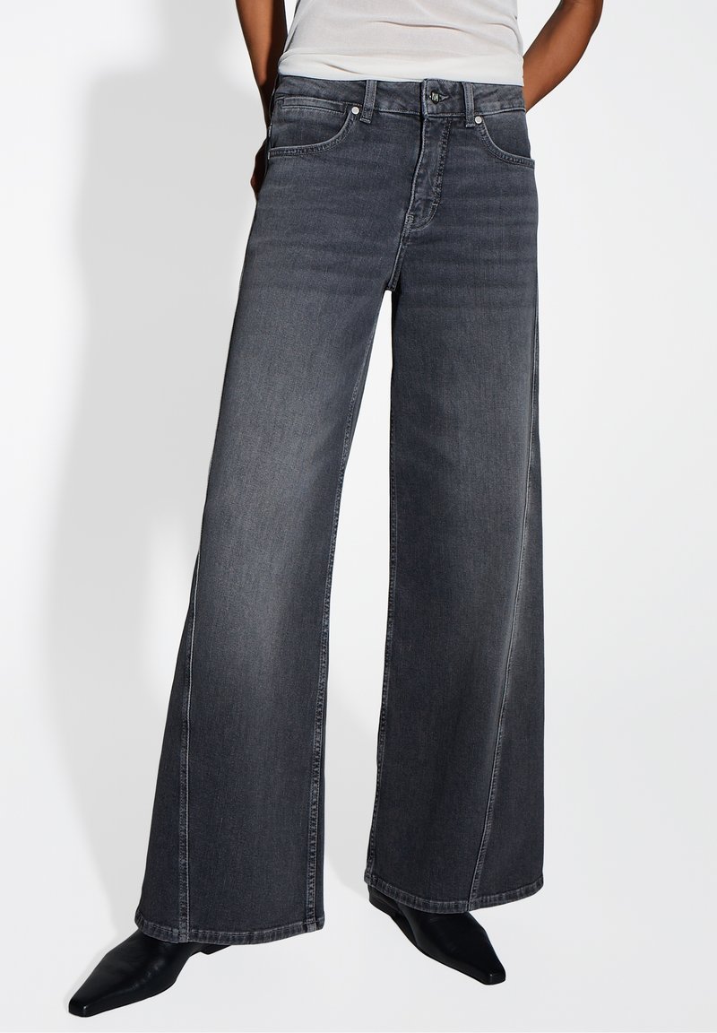 Personne portant un jean noir à jambes larges et des chaussures pointues noires, debout contre un fond clair uni.