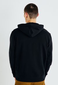 Felpa nera con cappuccio dal tocco morbido, con tasca a marsupio e polsini a costine, vista da dietro. Design semplice senza motivi.