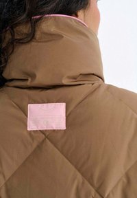 Brązowa pikowana kurtka z wysokim kołnierzem z różowym obszyciem i różową metką z tyłu, charakteryzująca się gładką fakturą i wzorem rombów.