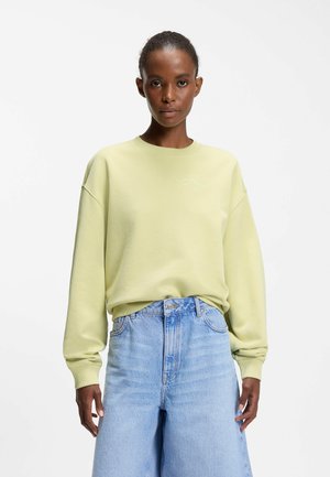 Frau trägt ein hellgelbes Sweatshirt mit kleinem weißen Logo auf der Brust und hochtaillierte, hellblaue, weit geschnittene Jeans, steht vor weißem Hintergrund.