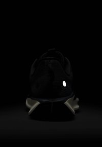 Zapatilla deportiva negra con parte superior de malla, detalles reflectantes y suela acolchada. Presenta un diseño elegante y un tacón pronunciado.