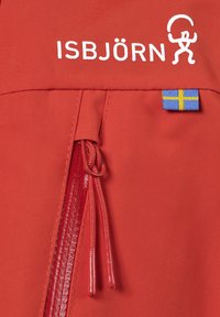 Röd tyg med slät textur har en dragkedjeficka, ISBJÖRN-logotyp och en liten svensk flagga som etikett.