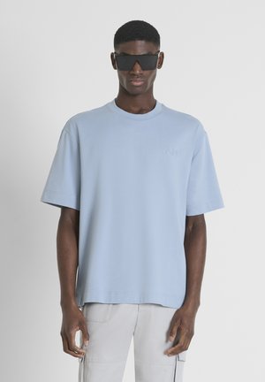 Lichtblauw oversized t-shirt met korte mouwen, een ronde halslijn en een subtiele geborduurde logo. Model draagt zwarte zonnebril en grijze broek.