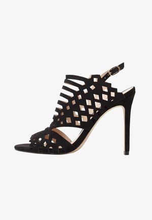 Sandalias de tacón alto de ante negro con patrones geométricos recortados, puntera abierta, tira trasera con hebilla y tacón de aguja delgado.