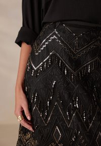 Black skirt with intricate geometric patterns. Embellished with silver and gold sequins, featuring fringe details. Soft textured fabric.