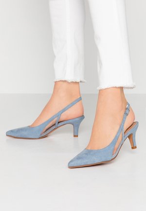 Pieds chaussés de escarpins slingback bleu clair en daim avec talons kitten, associés à un pantalon blanc court à l'ourlet effiloché, sur un sol blanc.