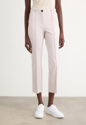 Pantalon court rose clair coupe slim avec coutures devant, bouton noir, fermeture éclair latérale, porté avec des baskets blanches sur une personne debout.