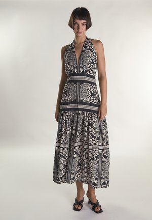 Mujer con un vestido sin mangas de tirantes halter con estampado floral en blanco y negro y falda de volantes, de pie frente a un fondo claro y liso.