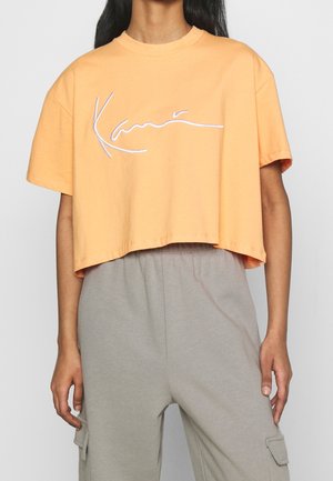 Person iført ferskenfarvet cropped T-shirt med hvid kursivlogo og lysegrå cargobukser, stående foran ensfarvet baggrund.