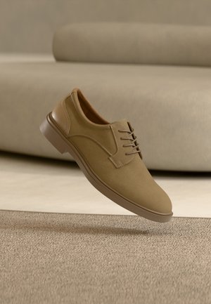 Chaussure décontractée beige à lacets flottant au-dessus d'un tapis texturé avec un canapé incurvé beige en arrière-plan.