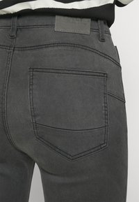 Jeans de denim gris con un diseño distintivo en el bolsillo trasero, detalles de costura sutiles y una etiqueta en la cinturilla. Se nota una textura suave.