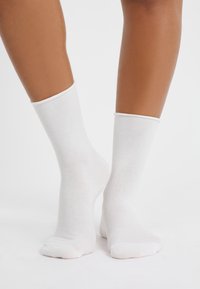 Calcetines de algodón blancos hasta el tobillo con puños acanalados, de textura suave y un diseño básico, descansando sobre piernas de color marrón claro contra un fondo neutro.