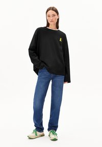 Frau in einem übergroßen schwarzen Langarmshirt mit einem gelben Logo, blauen Jeans und weißen Sneakers mit grünen Schnürsenkeln und Sohlen, stehend.