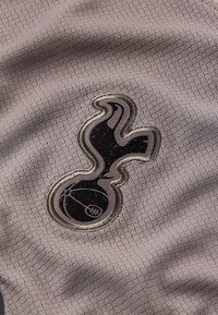 Tela gris con un patrón texturizado y un logo bordado en negro y plata que se asemeja a un pájaro sosteniendo un balón de baloncesto.