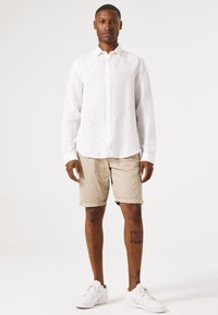 Chemise blanche en lin à manches longues, shorts beige retroussés, baskets blanches. Homme debout, les bras détendus, design simple et couleurs neutres.